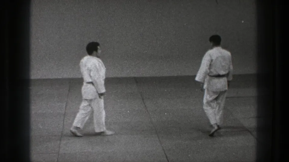Adult martial artists in white gi move across a dojo mat in grainy 8mm footage from Tokyo, 1971.