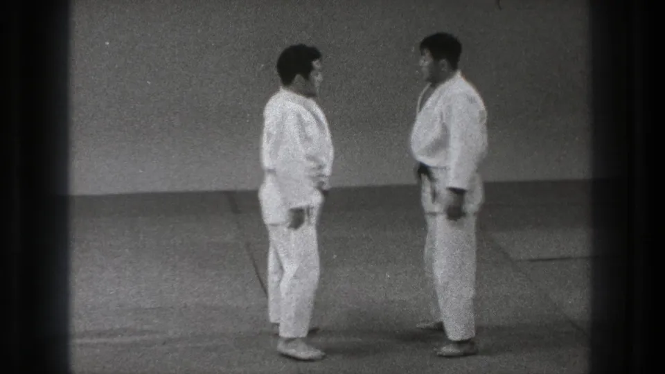 Grainy 8mm frame of two adult martial artists in white gi holding a close face-to-face stance in Tokyo, 1971.