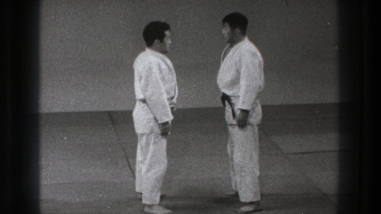 Two adult martial artists in white gi stand facing each other on floor mats.