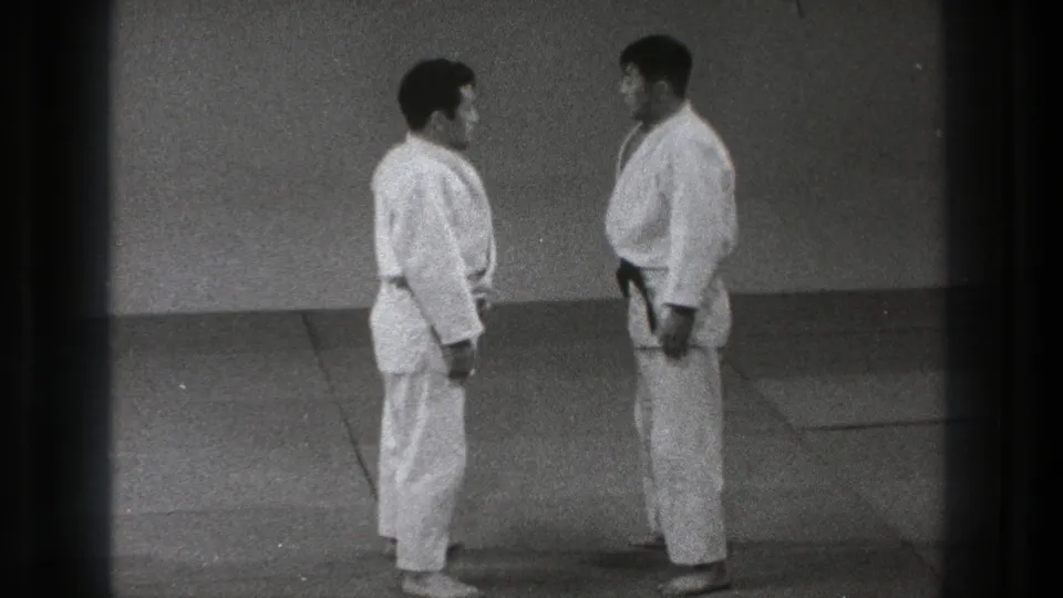 In a Tokyo 1971 martial arts setting, two adult practitioners in white gi and dark belts face each other before a grappling demonstration.