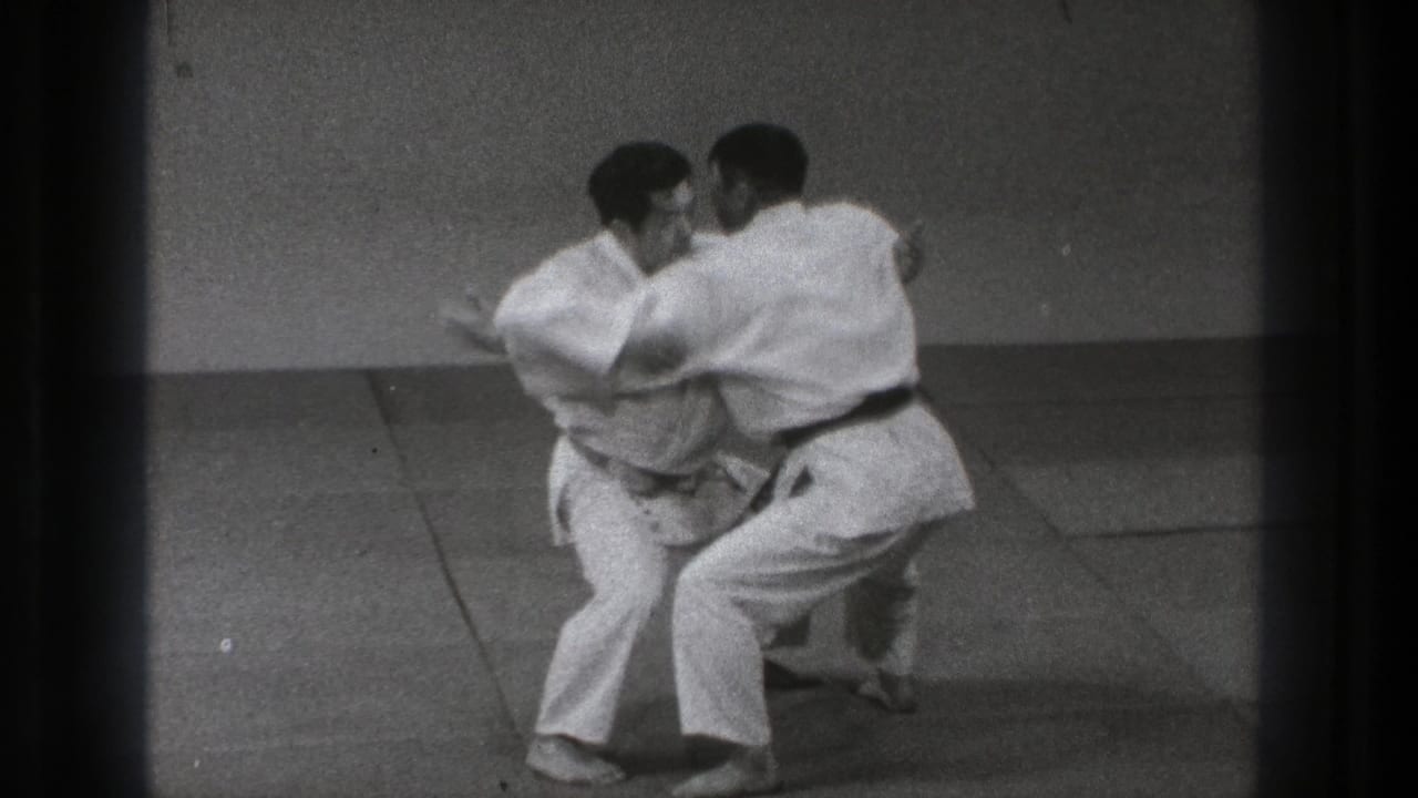 Two adult martial artists in Tokyo appear to begin a slow grappling move, with one stepping in close while both hold balance on the mats.