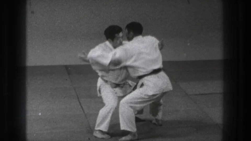 Two adult martial artists in Tokyo appear to begin a slow grappling move, with one stepping in close while both hold balance on the mats.