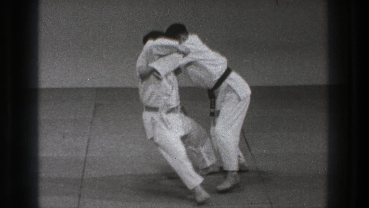In 1971 Tokyo 8mm footage, two adult men in white gi and dark belts grapple on a padded mat before a throw.