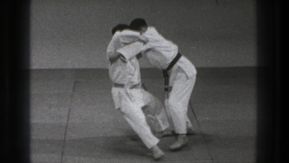 In 1971 Tokyo 8mm footage, two adult men in white gi and dark belts grapple on a padded mat before a throw.
