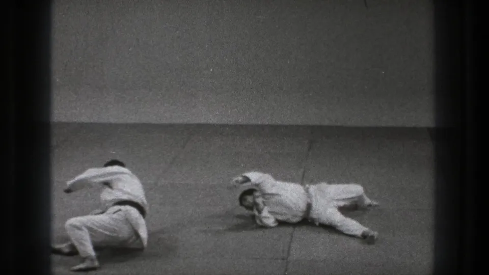 The throw resolves as one man drops backward and the other rolls or lands on the padded floor in the Tokyo martial arts sequence.