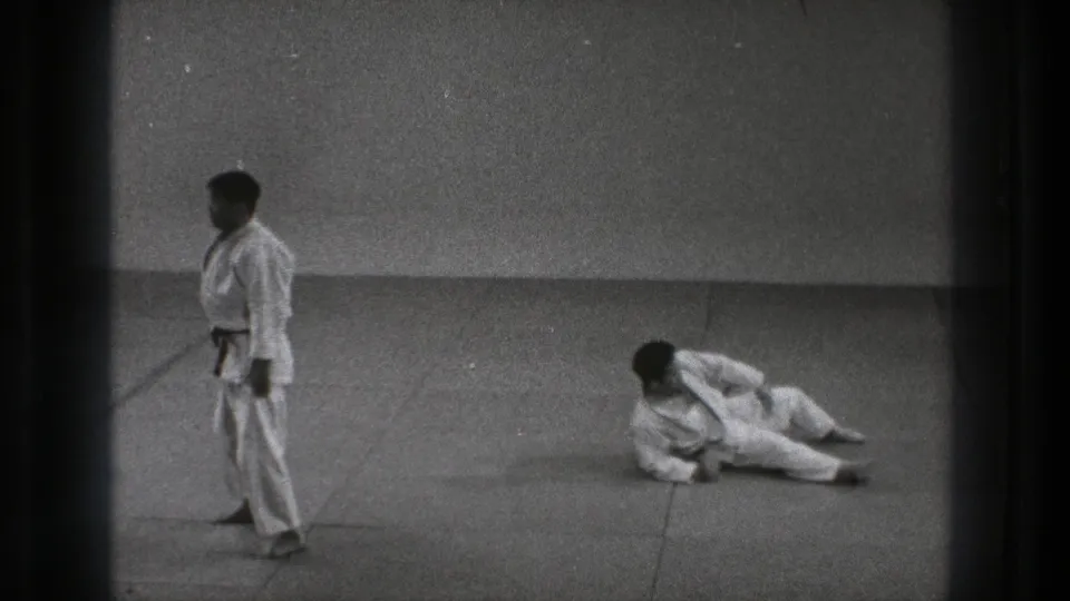 After the throw, one black-belt martial artist stands nearby while the other remains on his side on the padded floor.