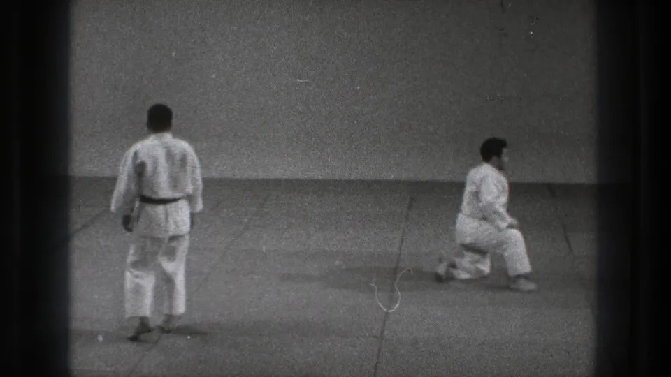 1971 Tokyo dojo footage showing two adult karate practitioners in white gi, with one standing and the other lowering into a stance.