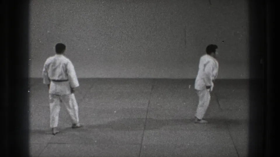1971 Tokyo karate practice scene with two adult men in white gi separated across a sparse indoor dojo floor.