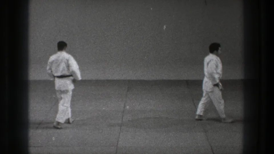 Black-and-white 8mm frame from 1971 Tokyo showing two adult karate practitioners moving across the dojo floor.