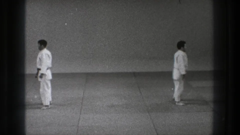 Two white-gi karate practitioners stand apart on a mat-like floor in black-and-white 1971 Tokyo archive footage.