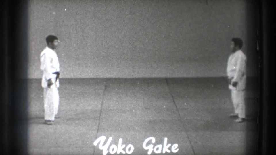 In 1971 Tokyo footage, two adult martial artists in white gi face each other across a plain mat as the handwritten title Yoko Gake appears below.