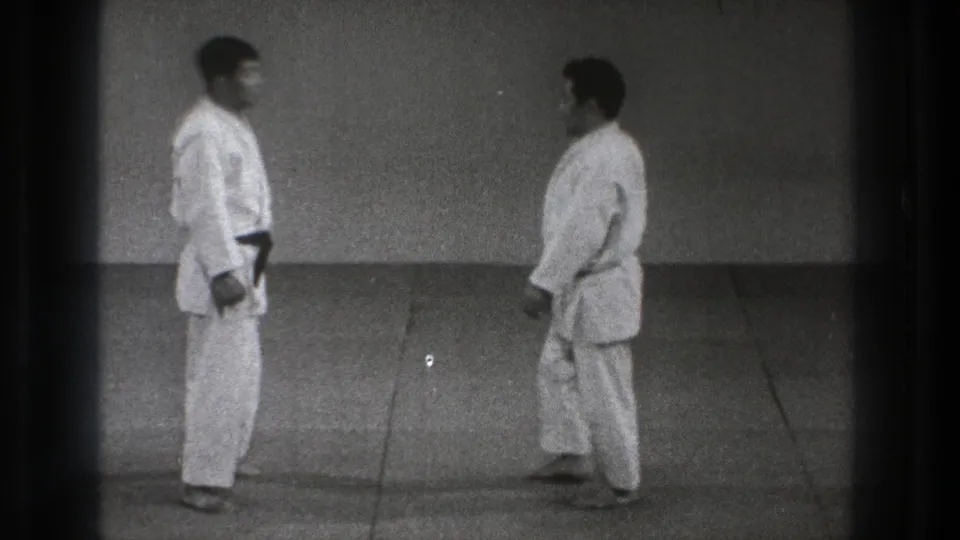 Two adult martial artists in white gi stand near each other on the mat, viewed in grainy black-and-white 8mm footage from Tokyo in 1971.