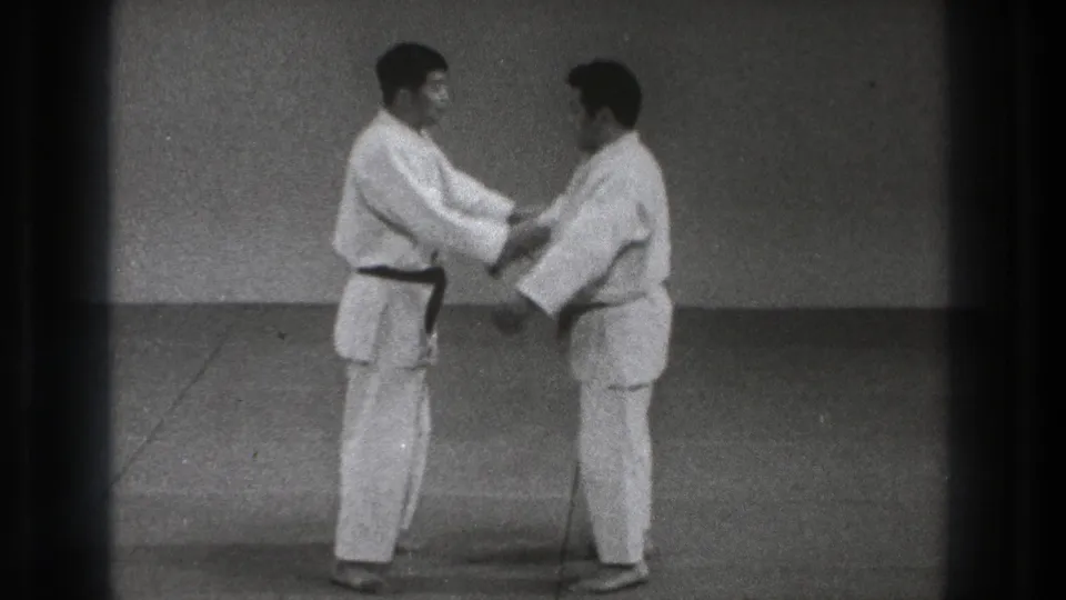 In the supplied 1971 Tokyo clip, two adult men in white gi stand on a plain mat in a dojo-like room while beginning a paired martial arts grip.