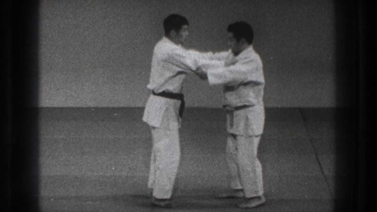 In this 1971 Tokyo martial arts frame, the dark-belted practitioner controls the other man's upper body as both stand on the mat in a sparse training room.