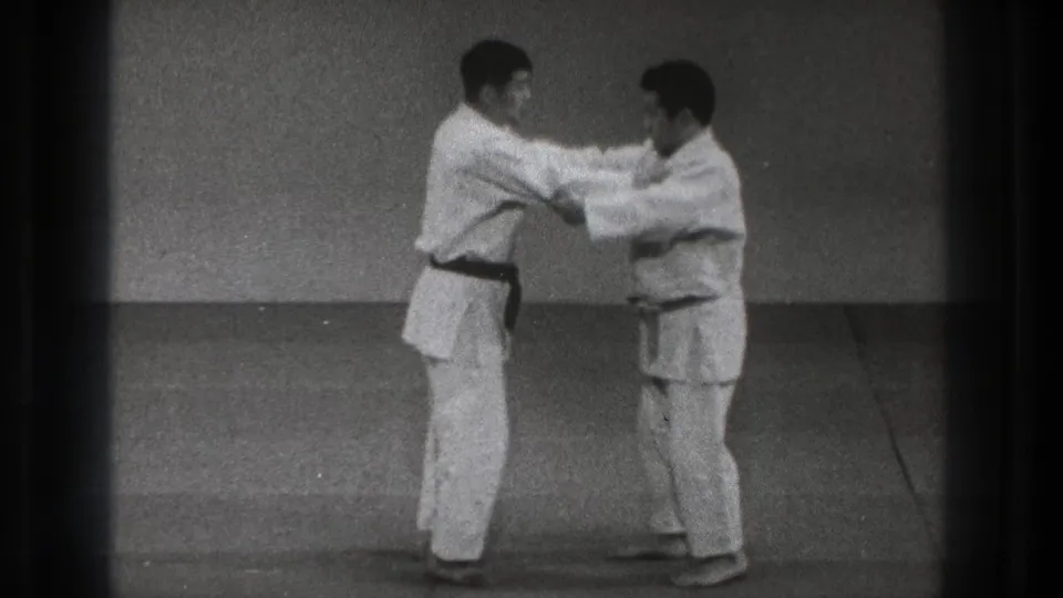 In this 1971 Tokyo martial arts frame, the dark-belted practitioner controls the other man's upper body as both stand on the mat in a sparse training room.