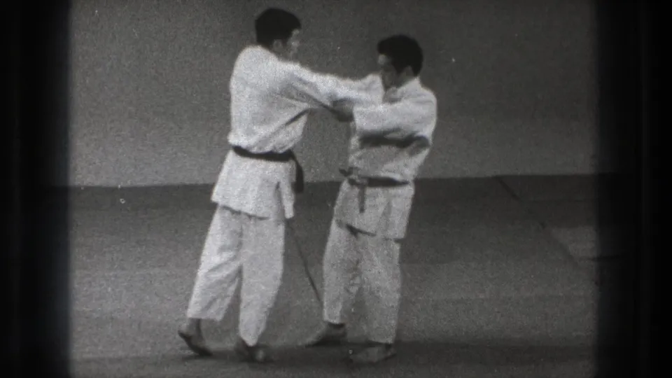 Near the end of the 1971 Tokyo clip, the dark-belted man maintains upper-body control while the other practitioner turns slightly during the paired dojo drill.