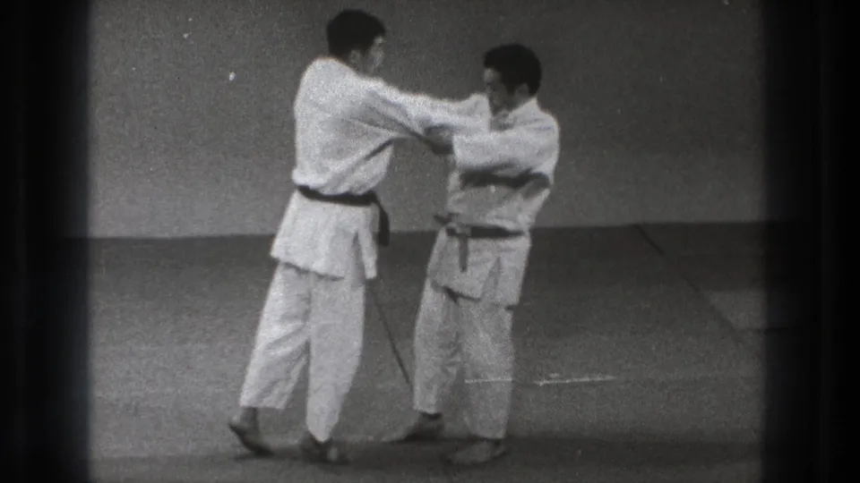 In Tokyo 1971 footage, two adult martial arts practitioners in white gi and dark belts begin a standing grip on a mat.