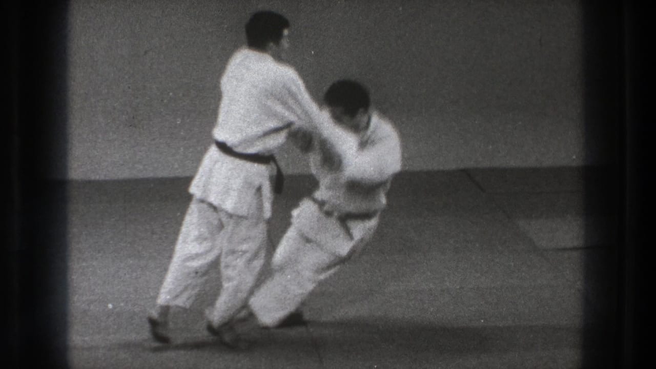 One adult practitioner controls his partner's arm as the partner drops toward the mat during a throw sequence in Tokyo 1971 footage.