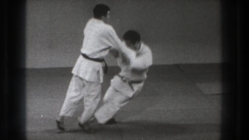 One adult practitioner controls his partner's arm as the partner drops toward the mat during a throw sequence in Tokyo 1971 footage.