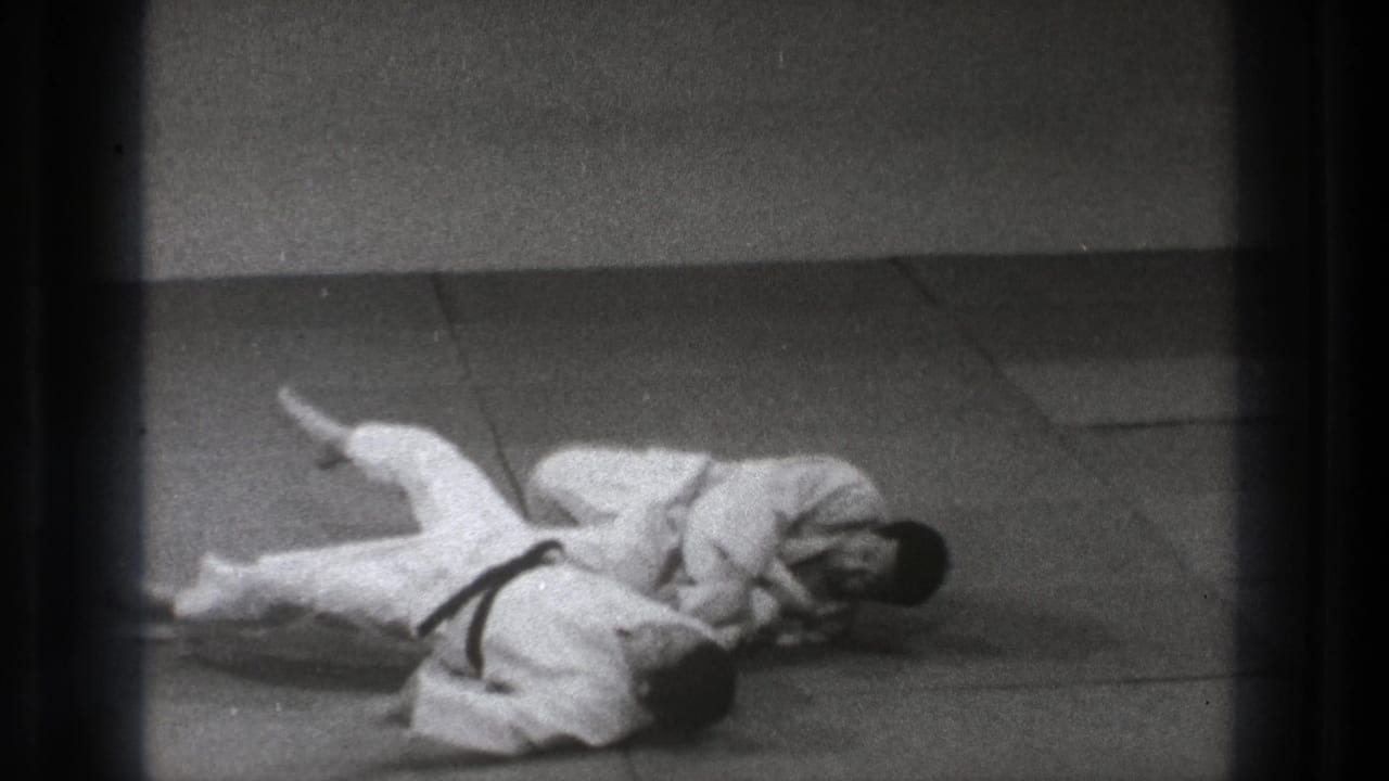 Two men in white gi grapple on a matted floor, one partly on top of the other.