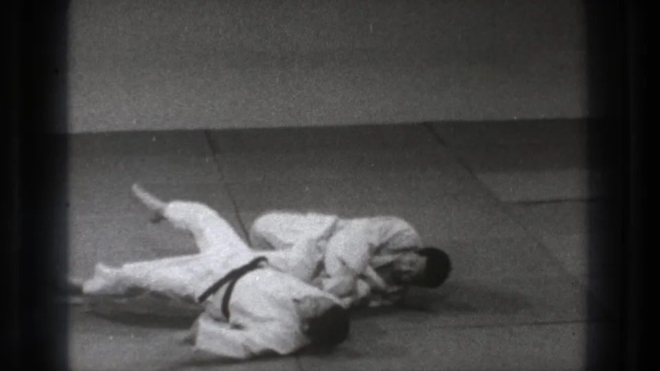 In 1971 Tokyo footage, two adult martial arts practitioners in white gi are down on a matted floor during a controlled grappling move.