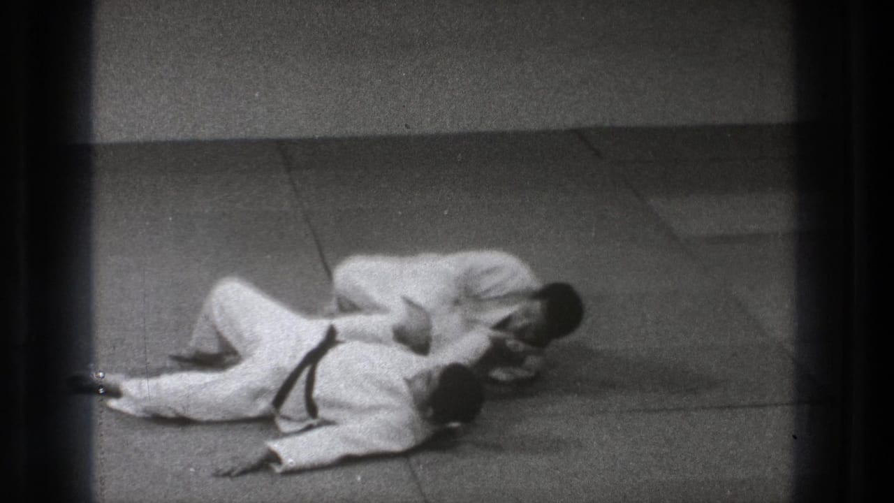 Two adult men in white gi remain on the tatami-like mat as one controls the other's upper body during a Tokyo martial arts practice sequence.