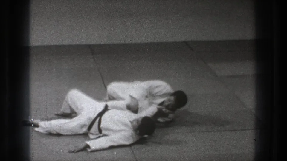 Two adult men in white gi remain on the tatami-like mat as one controls the other's upper body during a Tokyo martial arts practice sequence.