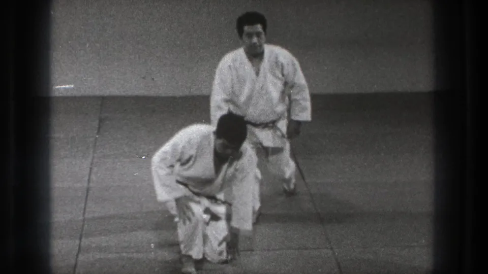At the end of the short sequence, two adult practitioners in white gi begin to rise from the mat in the Tokyo training space.