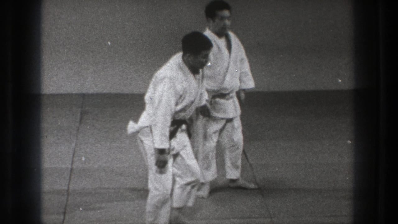 Two men in white gi stand on padded mats during judo practice.
