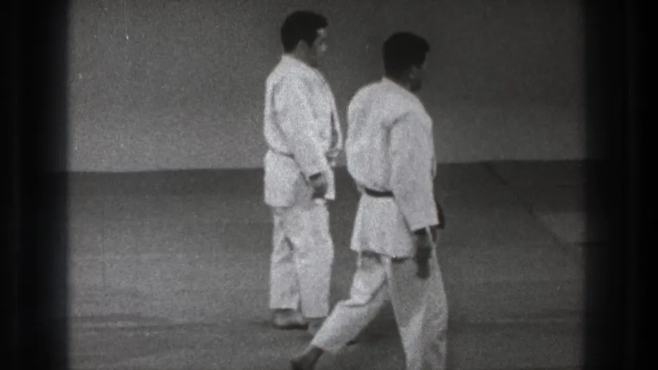 Two adult judo practitioners in white gi move across an indoor mat in 1971 Tokyo home-movie footage.