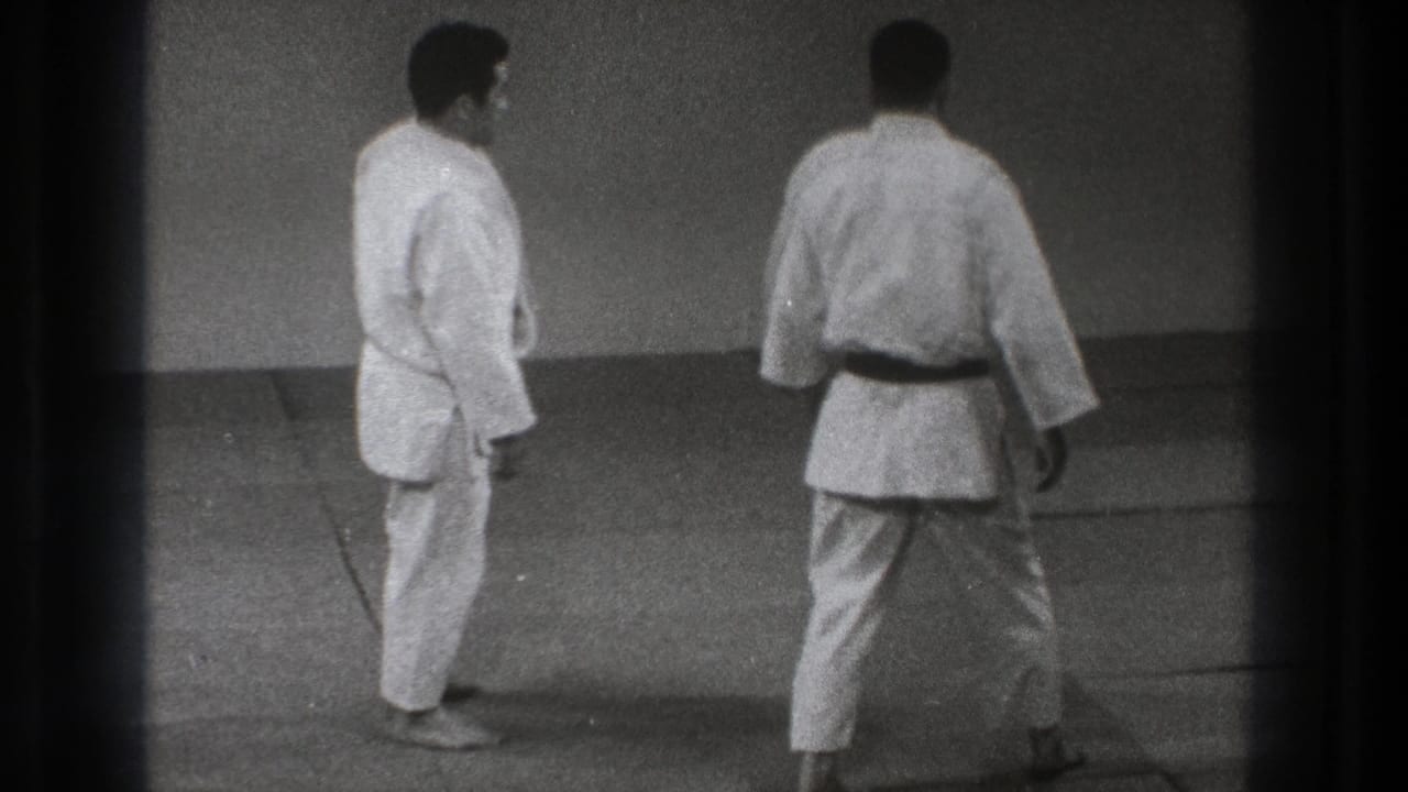 Two adult martial artists in gi pause on the mat, with one viewed from behind, in 1971 Tokyo judo practice footage.
