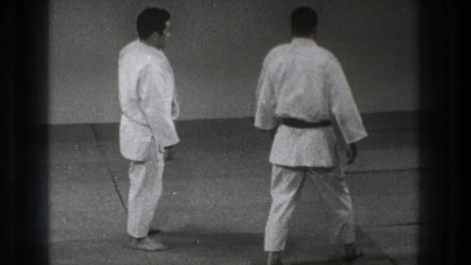 Two adult martial artists in gi pause on the mat, with one viewed from behind, in 1971 Tokyo judo practice footage.