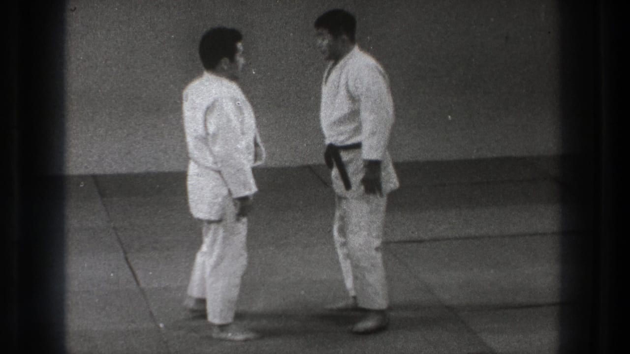 Two men in white karate gi stand facing each other on indoor mats.
