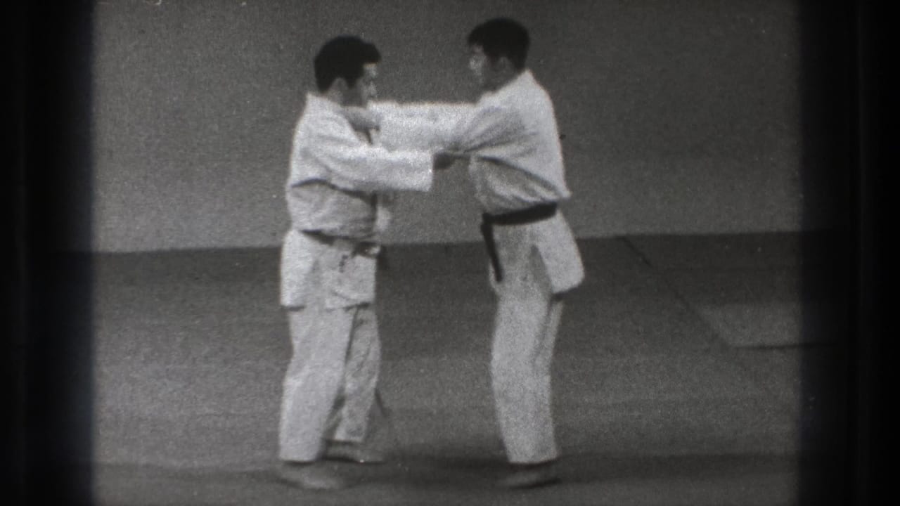 Two men in gi make arm contact in a standing karate technique on indoor mats in archival Tokyo footage.