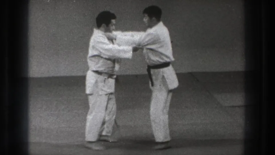 Two men in gi make arm contact in a standing karate technique on indoor mats in archival Tokyo footage.
