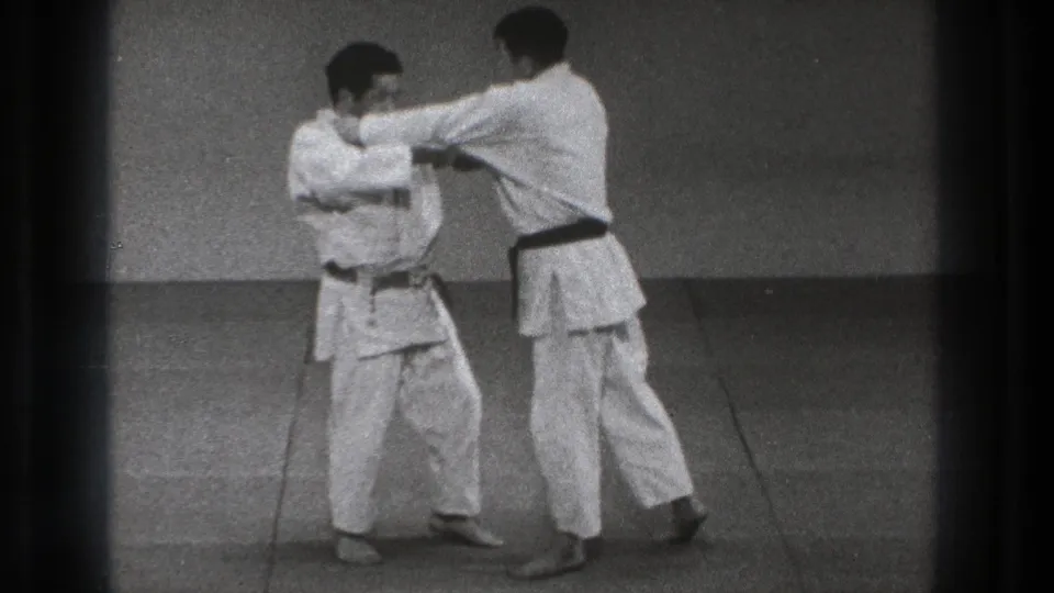 In Tokyo 1971 footage, two adult martial artists in white gi face each other on a mat, one wearing a dark belt.