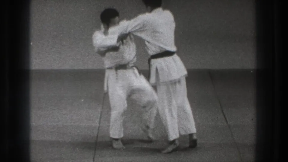 In the 1971 Tokyo judo sequence, one practitioner raises a leg while maintaining grips during the setup for a throw.