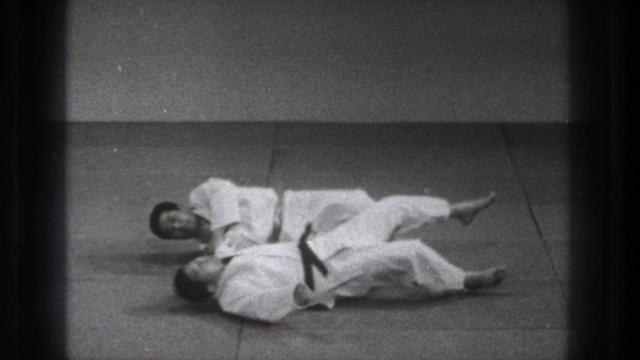 Two martial artists in white uniforms lie on floor mats during a ground technique.