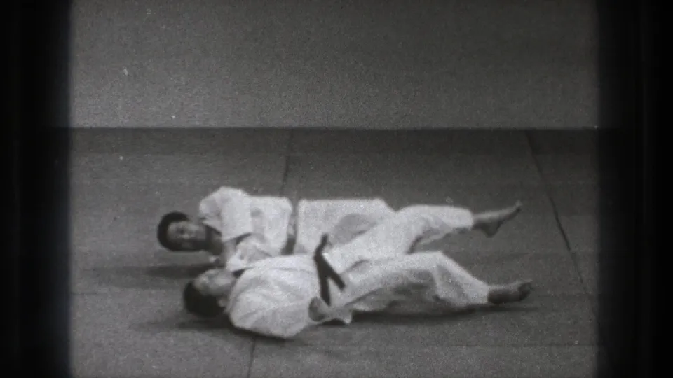 In the supplied 1971 Tokyo clip, two adult martial artists in white uniforms practice a grounded hold on plain indoor mats.
