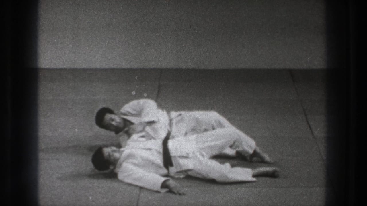 Two adult practitioners in white gi uniforms continue a controlled ground technique in a simple indoor dojo-like setting.