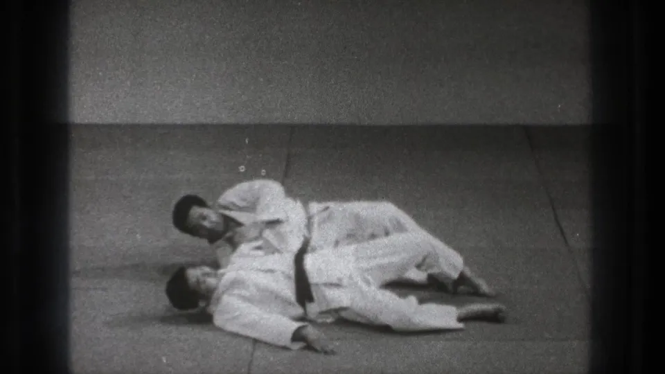 Two adult practitioners in white gi uniforms continue a controlled ground technique in a simple indoor dojo-like setting.