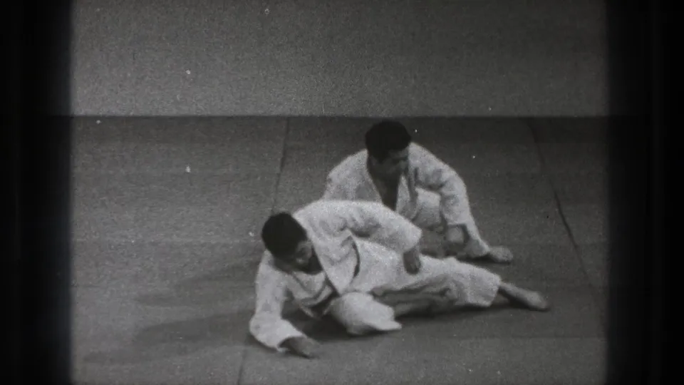The practitioners transition from a low hold, with one seated behind the other on the mat in the 1971 Tokyo training footage.