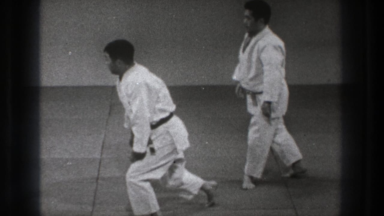 Two men in white gi on a matted floor, one stepping forward while the other stands nearby.