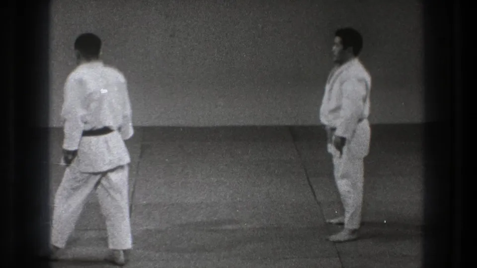 Two martial arts practitioners face off in a Tokyo practice setting, filmed on grainy 8mm in 1971.