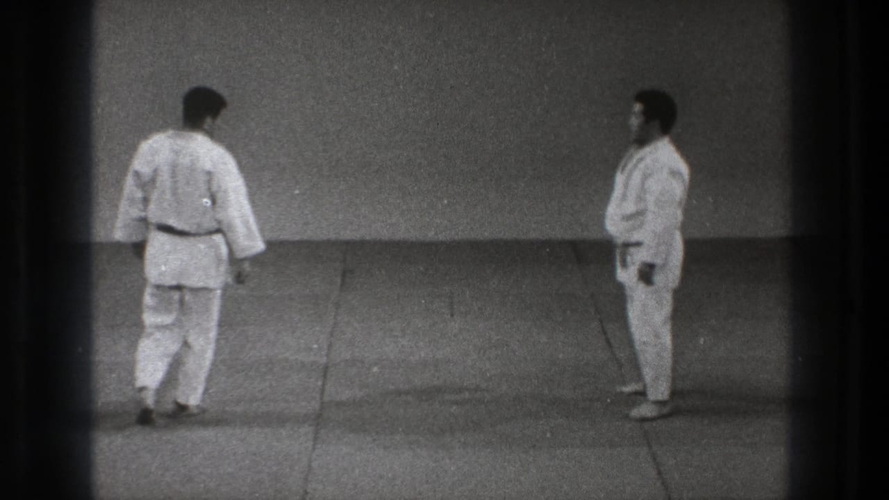 Grainy 1971 Tokyo frame of two martial arts practitioners standing apart on a plain matted practice floor.
