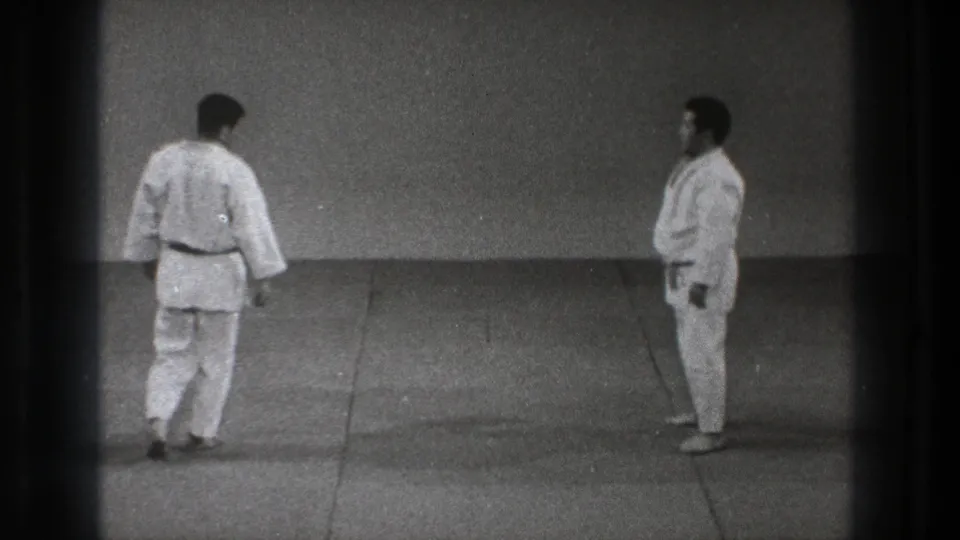 Grainy 1971 Tokyo frame of two martial arts practitioners standing apart on a plain matted practice floor.