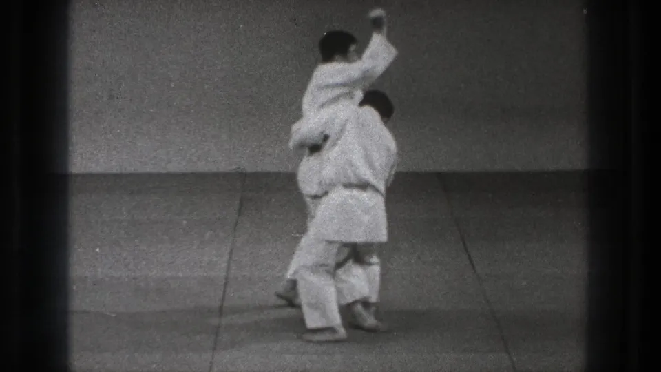 1971 Tokyo martial arts demonstration frame with one gi-clad practitioner raised over the defender during the technique.