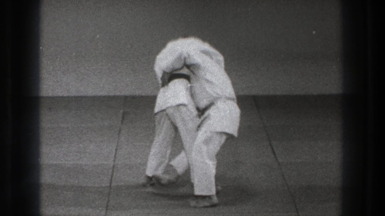 Two adults in white gi bend forward during a martial arts throw setup on a mat.