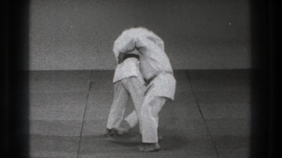 In 1971 Tokyo footage, two adults in white gi and dark belts lean into a close martial arts throw position on a plain mat.