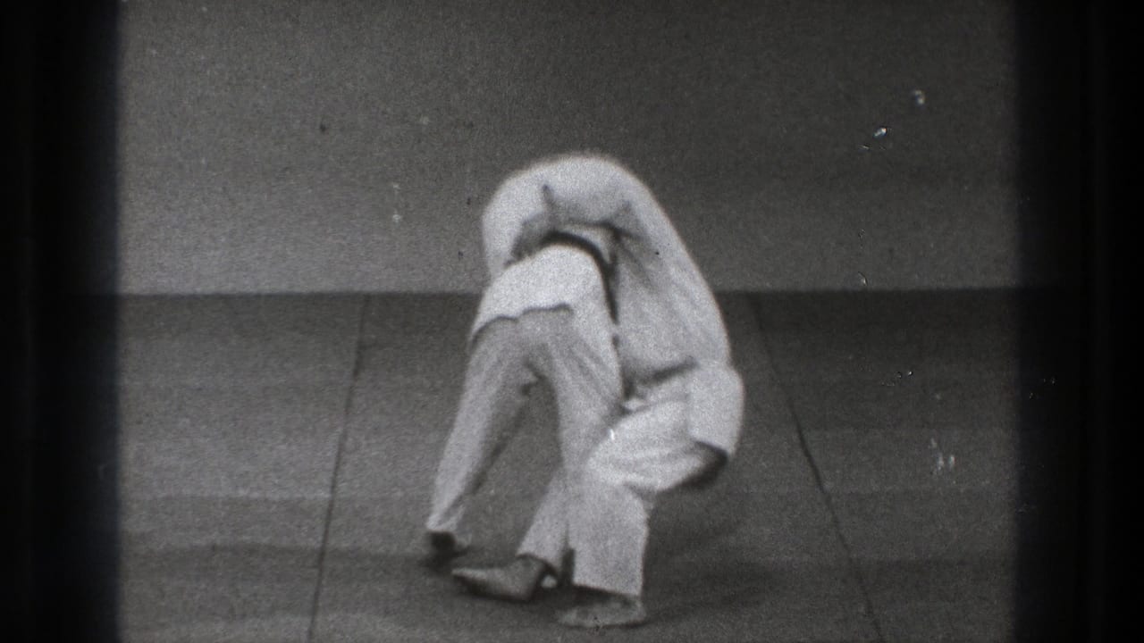 The Tokyo martial arts sequence shows one adult in a white gi lifting another into the early phase of a throw.